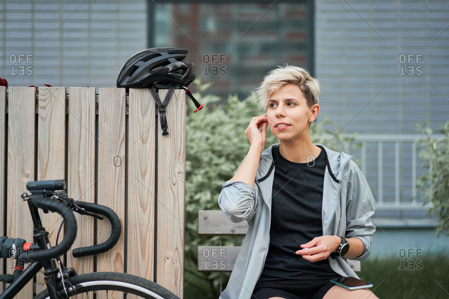 Female courier talking on phone while sitting on bench next to bicycle on background of building on summer day
