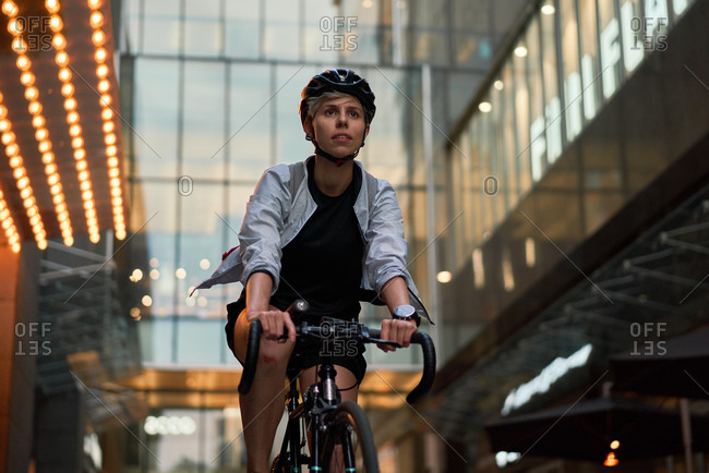 Woman courier cycling near tall building with glass windows in city on summer day