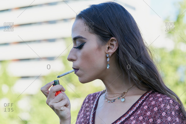 Young trendy ethnic female with makeup in accessories lightning cigarette with lighter in town