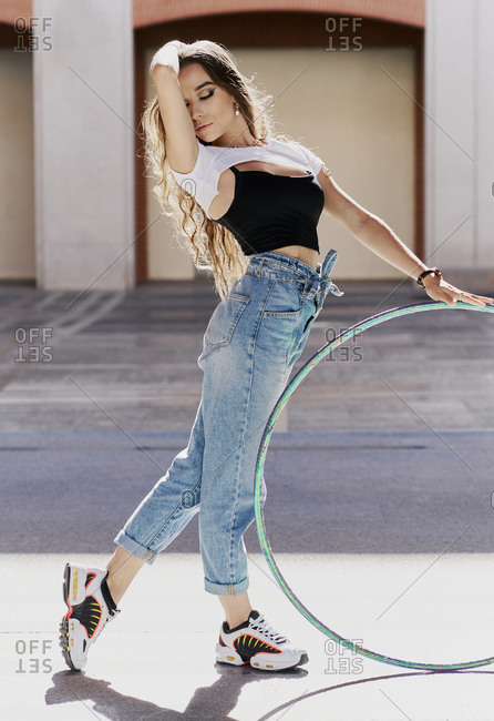 Full body side view of long haired slim female with eyes closed in trendy casual wear performing dance movement with hula hoop on paved square against urban building with columns
