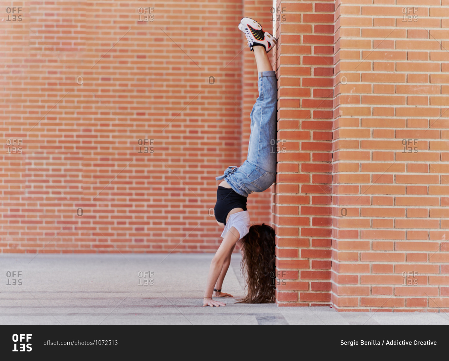 Full body side view of young female dancer in trendy casual outfit performing handstand near brick column on city street