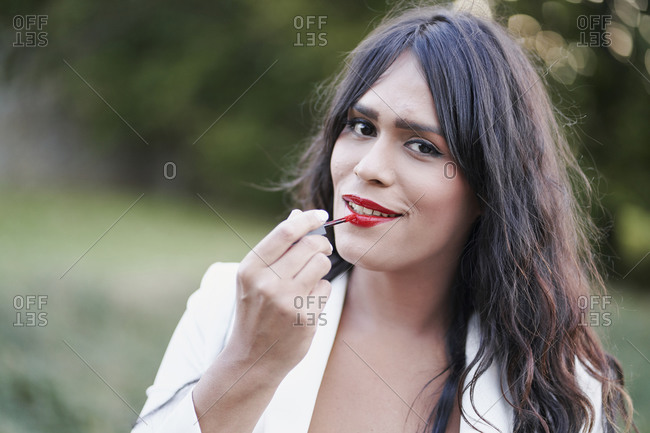 Charming female with long hair applying red lipstick while standing on street and smiling at camera