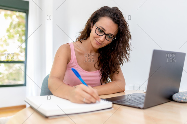 Positive young female student in casual clothes and eyeglasses sitting at table with laptop and writing information in copybook during online studies at home