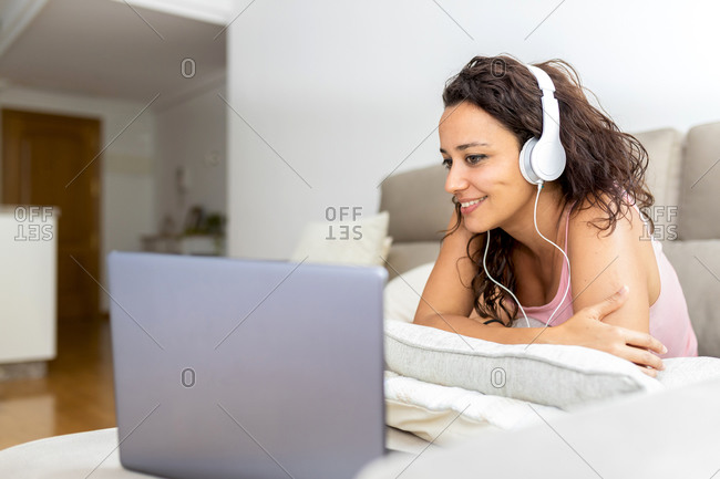 Cheerful relaxed female lying on sofa with laptop and enjoying movie in headphones while chilling at weekend at home