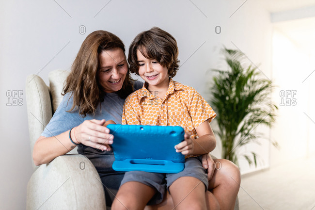 Cheerful kid with bright netbook sitting on couch with content mother in house