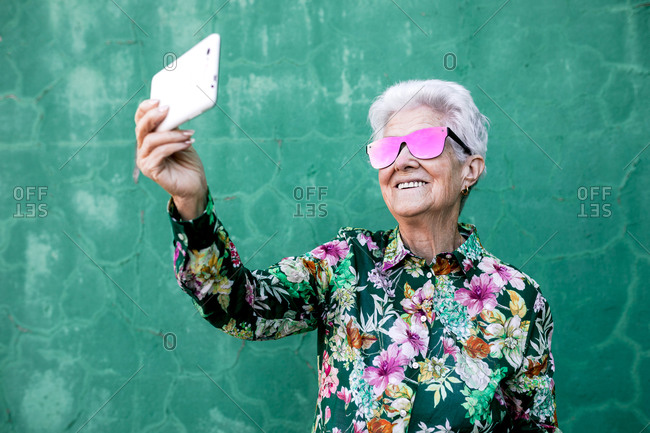 Happy aged gray haired female in stylish outfit and sunglasses taking selfie on mobile phone while standing against green wall