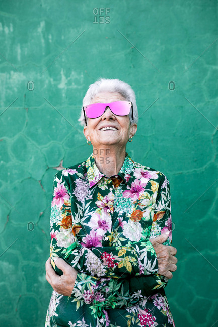 Confident senior gray haired female in stylish colorful clothes and trendy sunglasses smiling happily while standing against green wall
