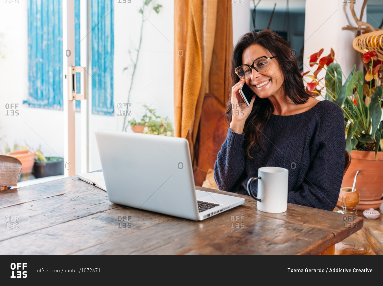 Positive adult ethnic female entrepreneur in casual wear talking on mobile phone and writing information in planner while standing near table with laptop and working remotely in cozy country house