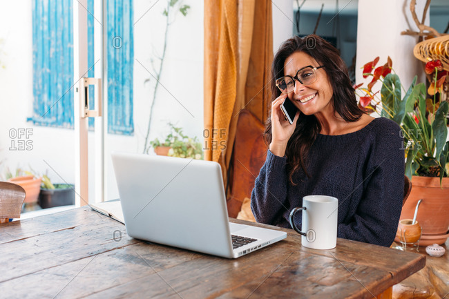 Positive adult ethnic female entrepreneur in casual wear talking on mobile phone and writing information in planner while standing near table with laptop and working remotely in cozy country house