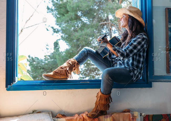 Side view of cheerful ethnic female dressed in cowboy style clothes with boots and hat sitting on windowsill and playing guitar while resting in rural house in summer day