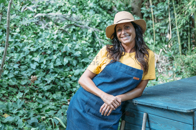 Positive adult ethnic female in denim apron and with hat in hand standing in tropical garden with green lush foliage in summer day