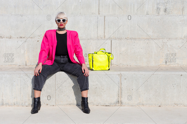 Confident female model wearing bright pink jacket sitting on stairs with vivid yellow handbag in city and looking at camera
