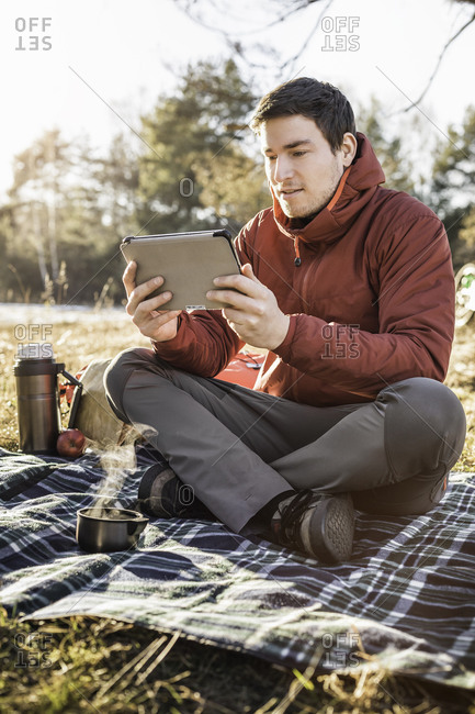 Young man sitting on picnic blanket in forest looking at digital tablet