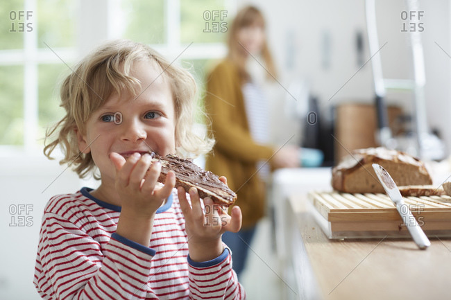 Young boy eating bread with chocolate spread, mother in background