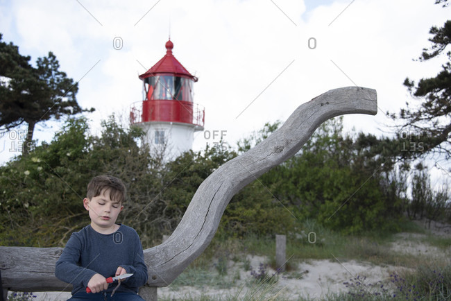 Germany, mecklenburg-west pomerania, hiddensee, boy carves in front of the gellen lighthouse, which stands on a grass dune. blue sky with clouds in the background, baltic sea.