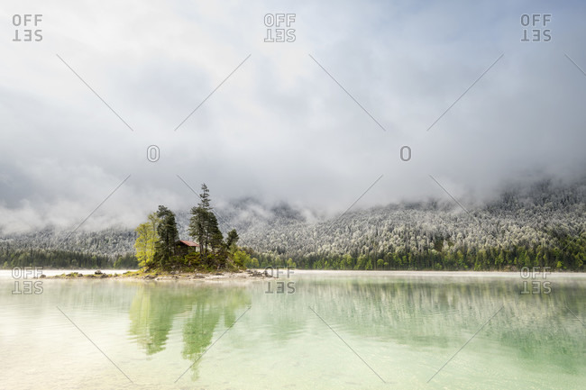 A small lonely island with a log cabin and red roof, in the middle of the beautiful eibsee below the zugspitze in the bavarian alps of the wetterstein mountains during the eisheiligenfresh snow in the background on the forest and cloud mood.