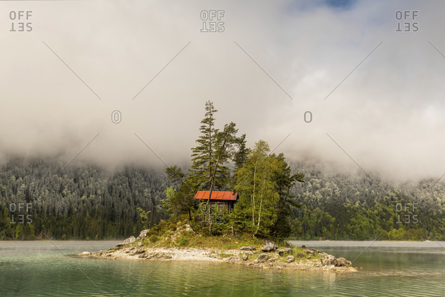 A small lonely island with a log cabin and red roof, in the middle of the beautiful eibsee below the zugspitze in the bavarian alps of the wetterstein mountains during the eisheiligenfresh snow in the background on the forest and cloud mood.