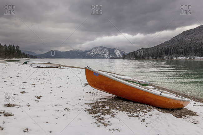Onset of winter on the banks of the walchensee in the bavarian prealps. in the foreground a rowboat and in the background the Herzog stand with snow and dark clouds in spring.