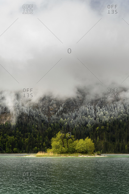 Small green island in the eibsee, below the zugspitze in the bavarian alps of the wetterstein mountains during the ice saints in spring. in the background snow on the fresh green of the forest and thick rain clouds.