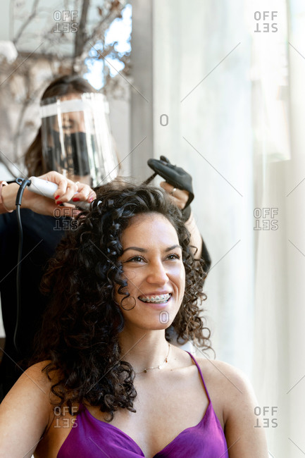 Professional hairdresser in black protective mask using curling tongs while making hairdo to long haired female customer in salon