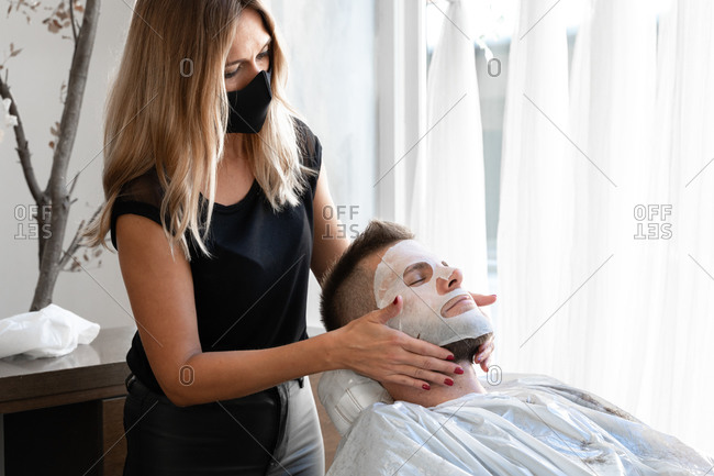 Professional female master in protective mask applying moisturizing facial mask to relaxed bearded male customer during grooming procedure in salon