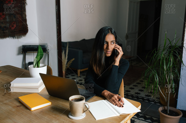 Busy ethnic female entrepreneur sitting at table in home office and speaking on cellphone about new startup project while working remotely and looking away