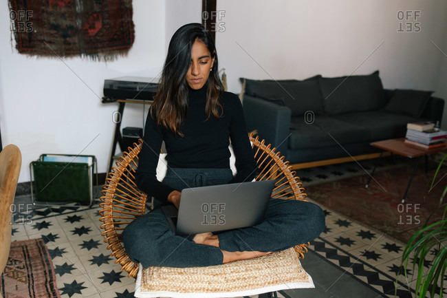 Serious Indian female freelancer sitting with crossed legs in wicker chair and typing on netbook while working online from home