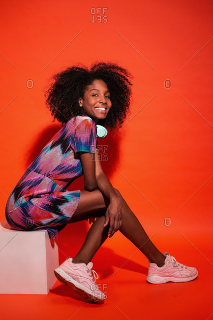 Full body side view of black woman with Afro hairstyle wearing vivid dress with pink sneakers sitting on red background