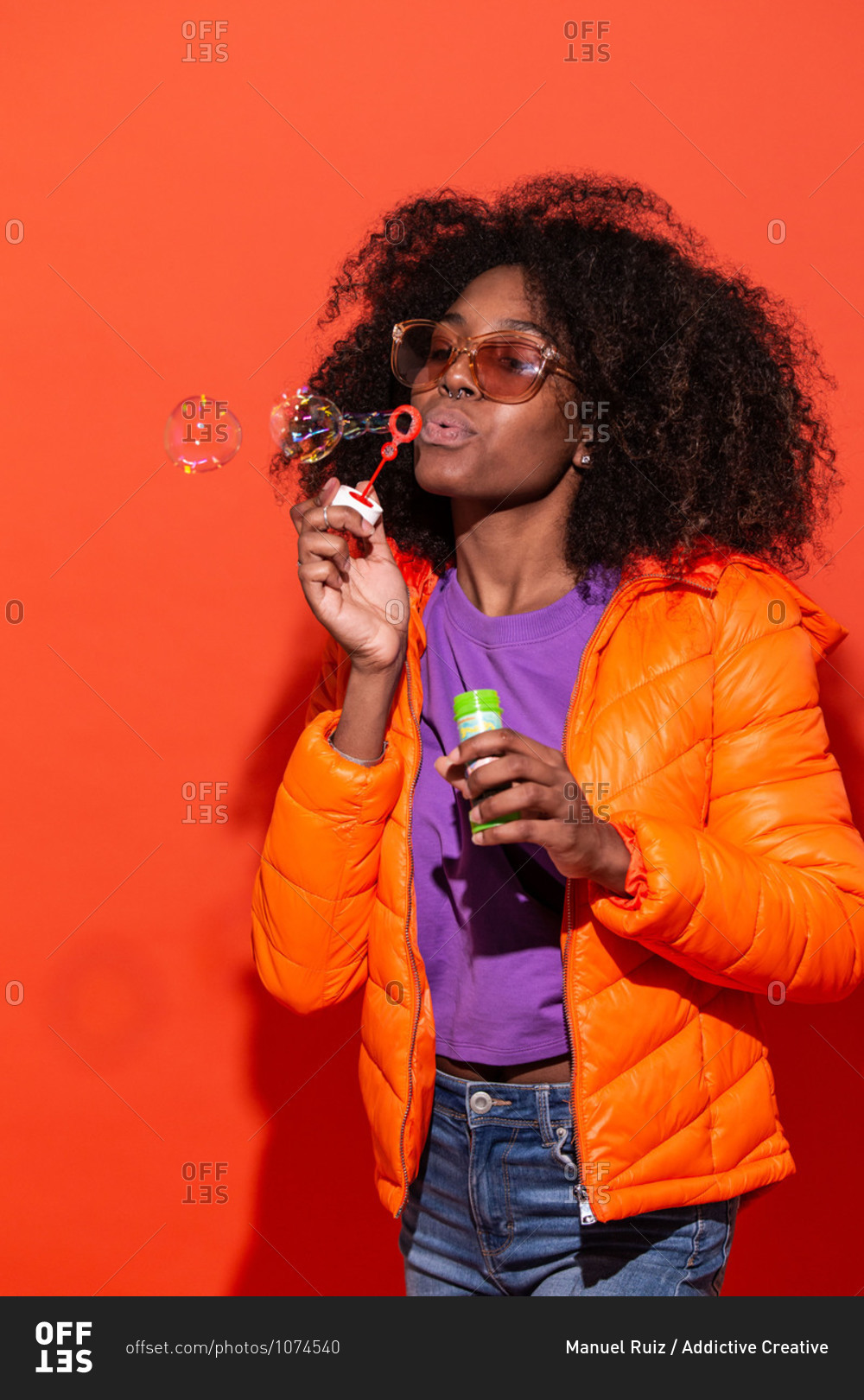 Modern African American female in colorful jacket blowing soap bubbles having fun in studio on red background