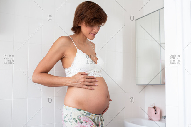 Side view of peaceful pregnant female in bra standing in bright bathroom and tenderly touching tummy while looking down