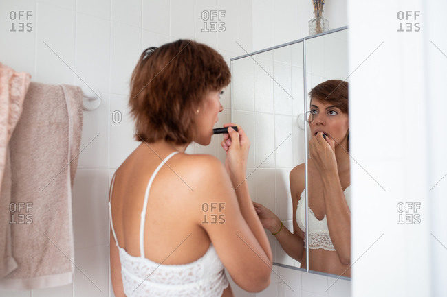 Back view of focused female in bra standing in bathroom and applying lip balm while doing makeup and looking in mirror