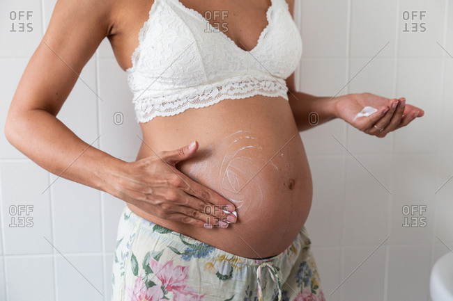 Crop anonymous pregnant female in bra standing in bathroom and applying moisturizing cream on tummy while doing daily skin care routine