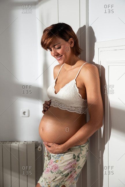 Smiling pregnant female in bra standing in white room and gently caressing tummy while looking down
