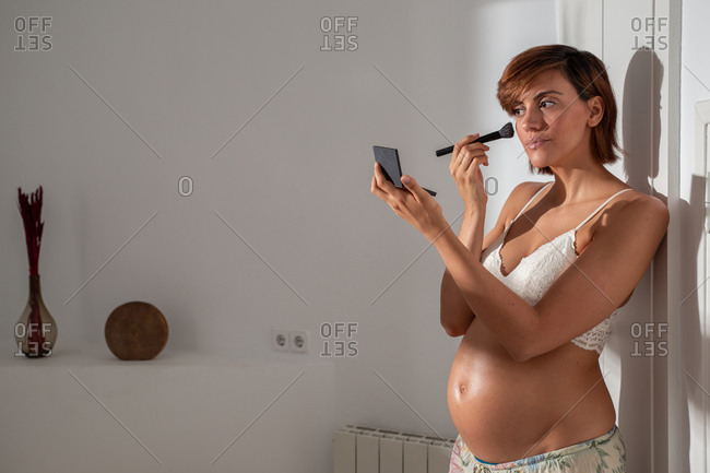 Charming pregnant female in bra standing in room and applying powder with brush while doing makeup
