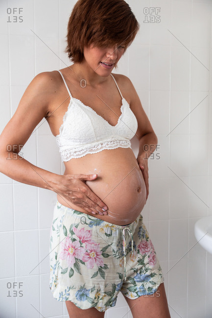 Side view of peaceful pregnant female in bra standing in bright bathroom and tenderly touching tummy while looking down