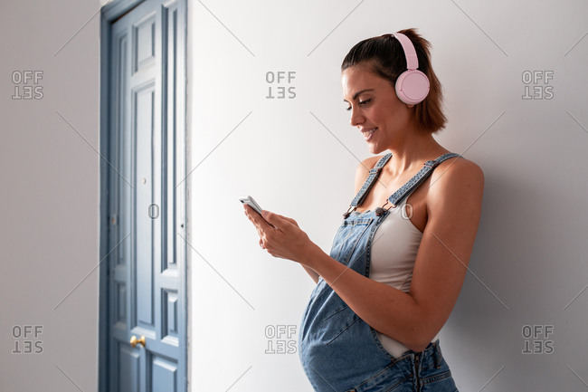 Side view of positive pregnant female in overalls standing in room and listening to music in headphones while choosing songs from playlist on smartphone