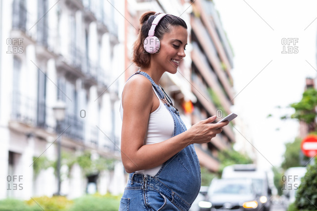 Side view of glad pregnant female in headphones standing on the street browsing on smartphone during city stroll in summer