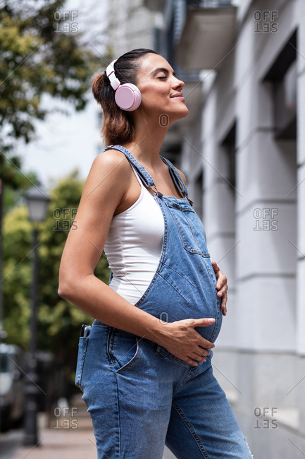 Side view of dreamy pregnant female in overalls and wireless headphones standing on street while enjoying songs and touching belly with closed eyes