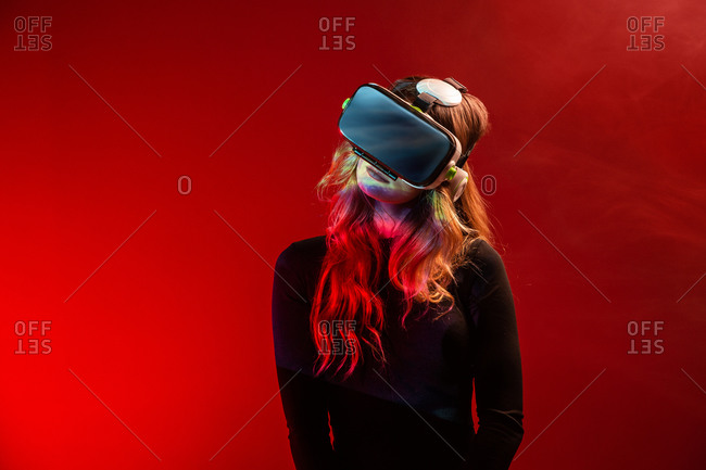 Side view of young unrecognizable blonde woman standing wearing virtual reality headset on colorful red studio background