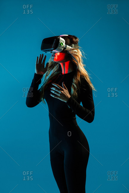 Side view of young unrecognizable blonde woman standing touching air and wearing virtual reality headset on colorful blue studio background