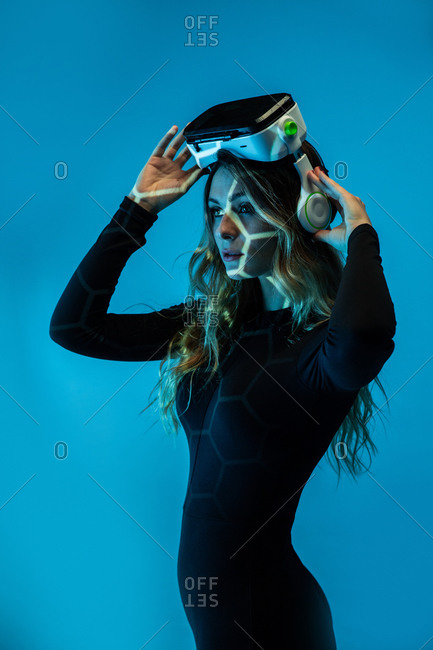 Side view of young blonde woman wearing virtual reality headset standing on colorful blue neon illumination studio background looking away