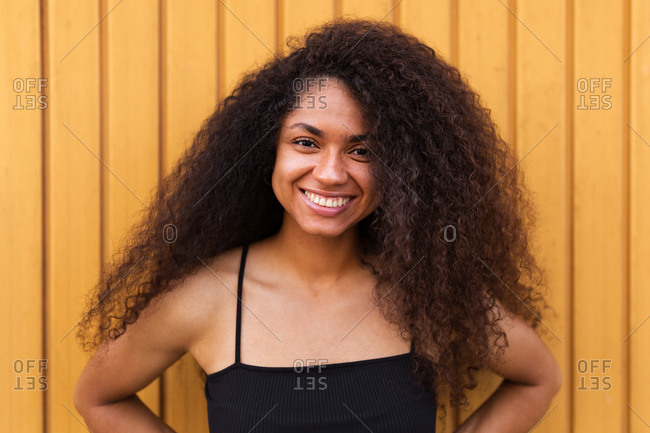 Delighted African American female with Afro hairstyle standing near wooden wall and sincerely smiling at camera