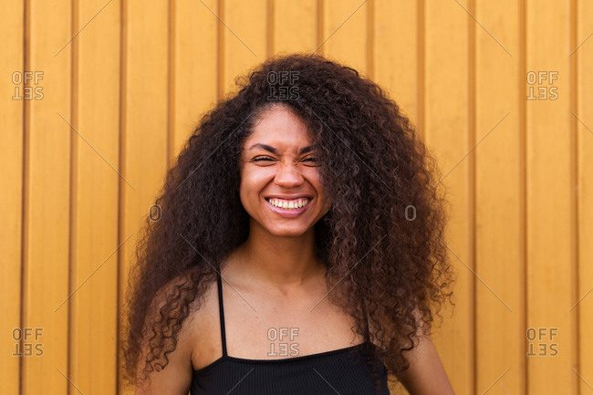 Delighted African American female with Afro hairstyle standing near wooden wall and sincerely smiling at camera