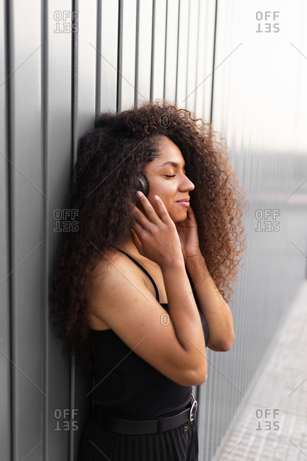 Lovely African American female with long curly hair and headphones in city and closed eyes