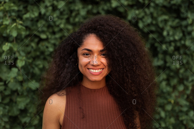 Optimistic African American young female with curly hair and charming smile looking at camera while standing in park against green plants