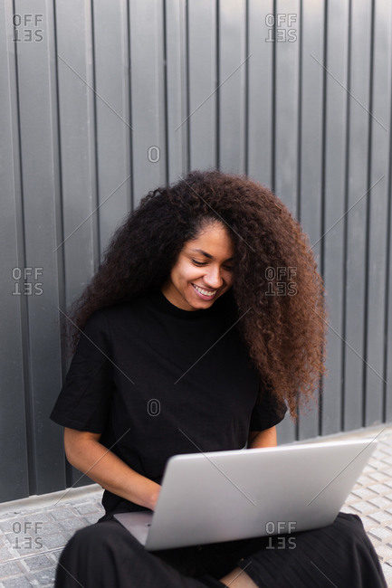 Cheerful African American self employed woman sitting on street with laptop