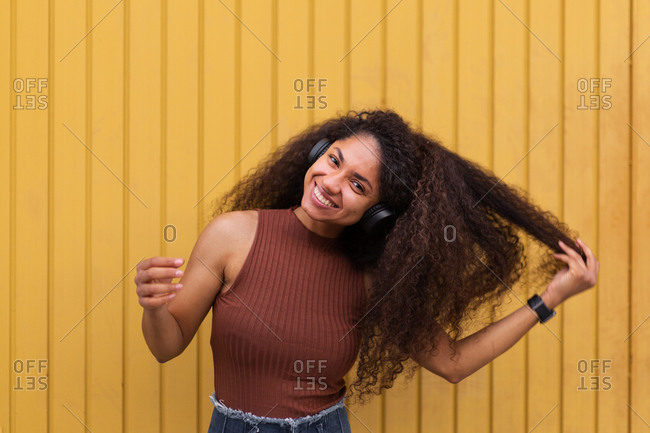 Lovely African American female with long curly hair and headphones in city and looking at camera