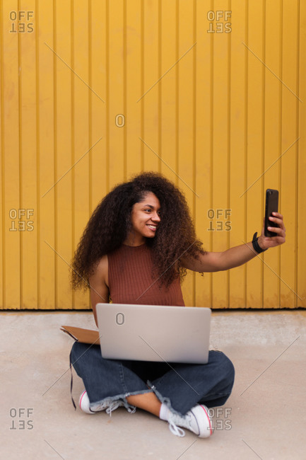 Cheerful African American self employed woman sitting on street with laptop and taking selfie on smartphone