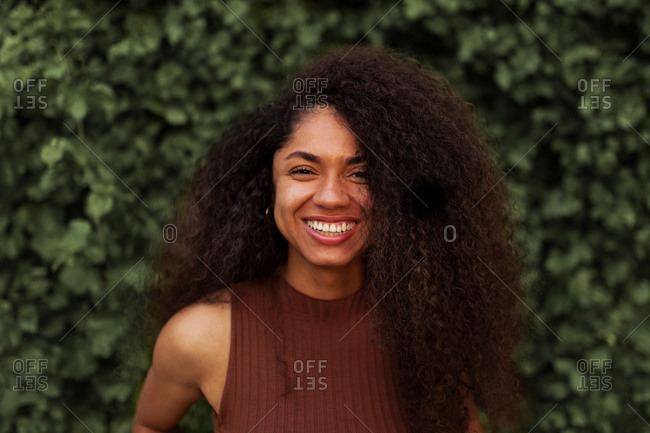 Optimistic African American young female with curly hair and charming smile looking at camera while standing in park against green plants