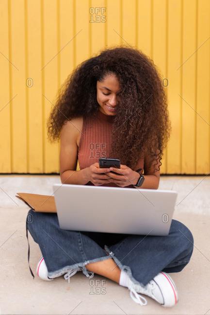 Cheerful African American self employed woman sitting on street with laptop and browsing smartphone